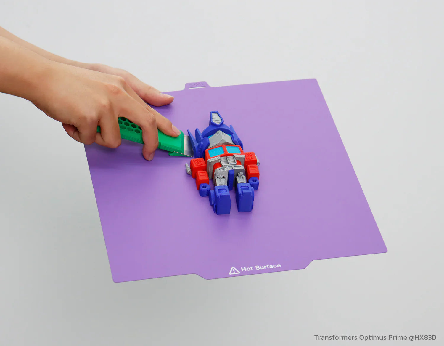 A hand lifts a red, white, and blue Optimus Prime-style toy from a purple 3D printer build plate using a green tool